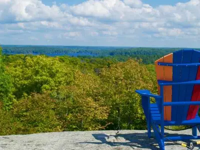A Muskoka Chair on a rocky outcropping overlooking trees with a lake in the background